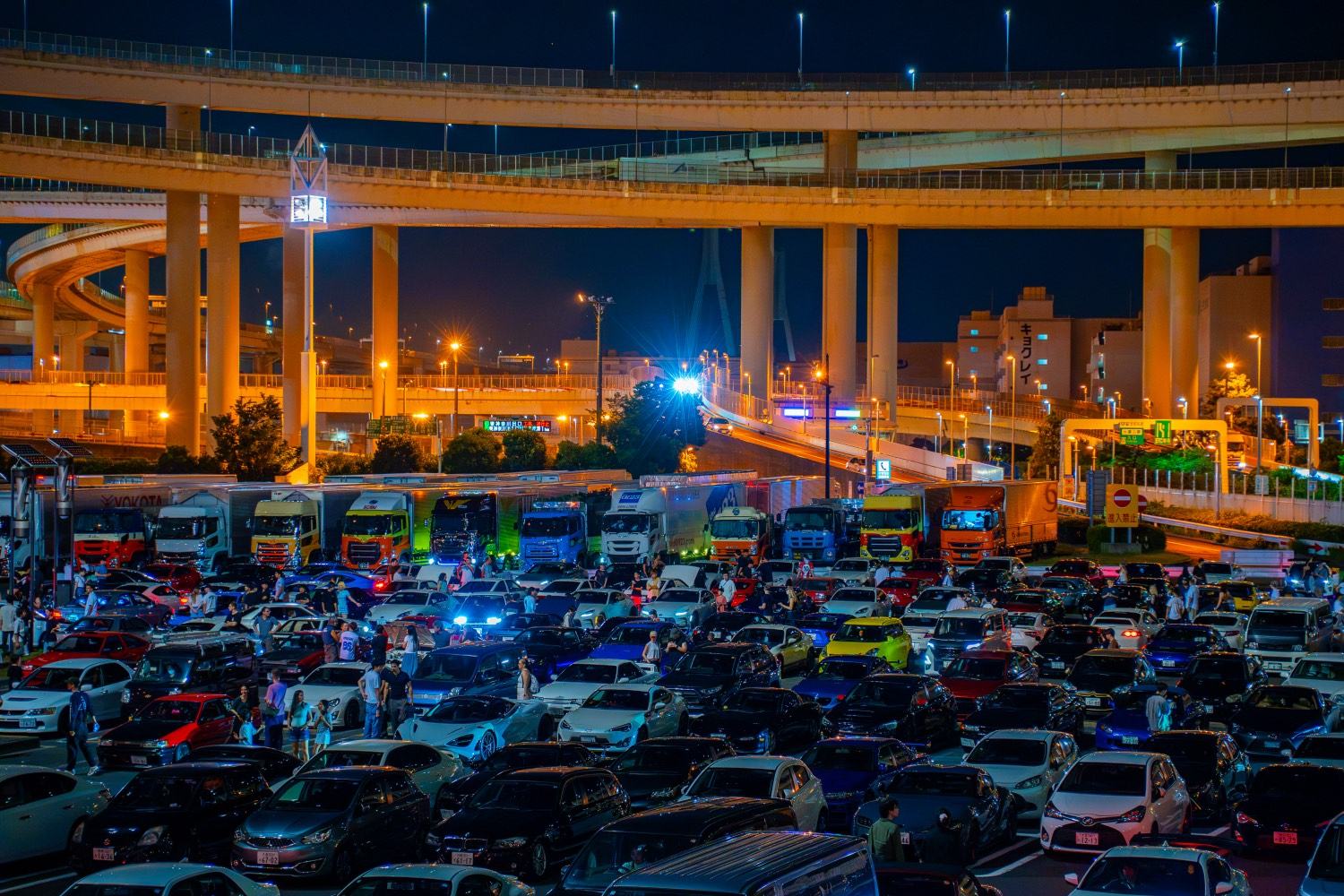 Daikoku Parking Area at night, several hundred cars filling the lot beneath the illuminated circular expressway ramp