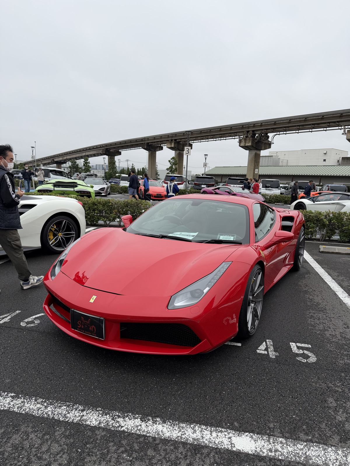 Red Ferrari 488 GTB parked at Daikoku Parking Area near Yokohama, with the elevated expressway loop overhead and other supercars gathered around it
