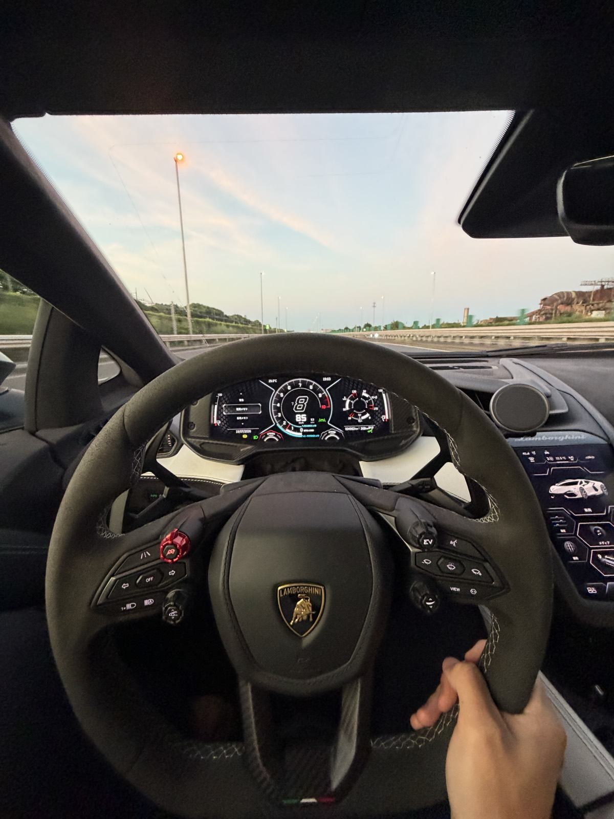 View from the passenger seat of the Lamborghini Revuelto on a Tokyo bayside expressway at dusk, with the driver's hand on the wheel and the digital dash lit